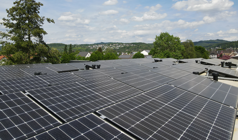 Photovoltaikanlage auf einem Flachdach mit zahlreichen nebeneinander montierten Solarmodulen in schwarzem Design. Im Hintergrund ist eine grüne Hügellandschaft mit verstreut liegenden Häusern und Bäumen zu sehen. Der Himmel ist bewölkt.