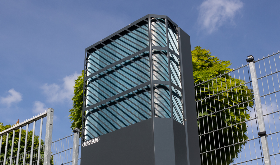LAMBDA Wärmepumpe der Firma ZEWOTHERM in anthrazitfarbener Ausführung, installiert im Außenbereich neben einem Metallzaun. Die Luft-Wasser-Wärmepumpe ist vertikal ausgerichtet und hebt sich vor einem sonnigen, blauen Himmel und einem grün belaubten Baum ab.