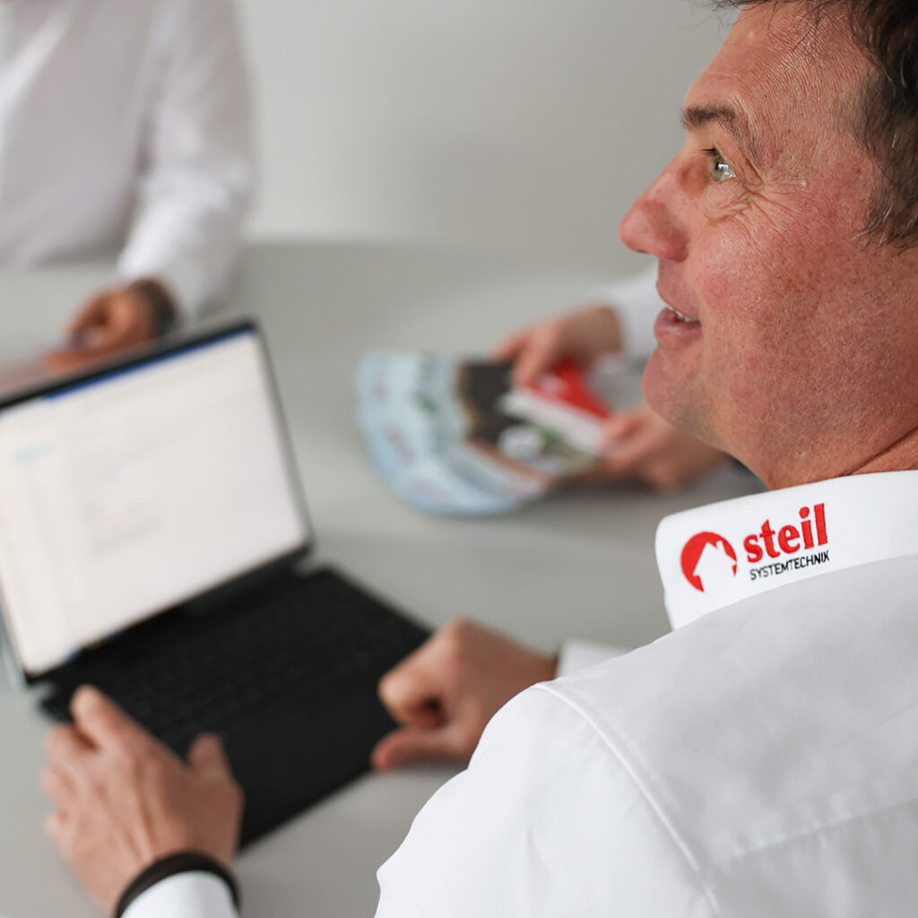 Eine Über-die-Schulter Aufnahme eines Steil Mitarbeiters der an einem Tisch sitzt und an seinem Laptop arbeitet. Er trägt ein weißes Hemd, auf dem Kragen ist gut sichtbar das rote Steil Logo zu sehen.
