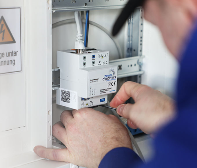 Eine Person in blauer Arbeitskleidung installiert oder wartet ein weißes EMS-Modul der Firma ZEWOTHERM mit der Aufschrift „zewo dynamics“. Das Modul ist per Kabel angeschlossen. Links im Bild befindet sich ein Warnschild mit einem Blitzsymbol und der Aufschrift „unter Spannung“.